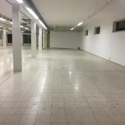 Przestronne, puste pomieszczenie przemysłowe z białymi ścianami, kwadratową podłogą z płytek, betonowymi kolumnami i rzędami świetlówek pod sufitem. Widoczne okna w górnej części ściany po prawej...