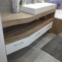 Nowoczesna łazienka z drewnianym blatem pod umywalką, białymi szufladami z minimalistycznymi uchwytami i pasującą obudową wanny, szara płytka podłogowa i dywanik.