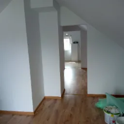 Poddasze po odświeżeniu, białe ściany w skosach, brązowe panele podłogowe, w tle widok na kolejne pomieszczenie, na podłodze wiadro z farbą i folia ochronna.
