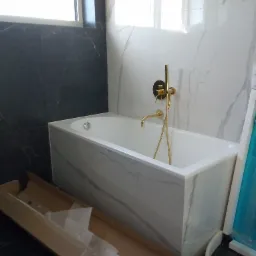 Nowoczesna wanna obudowana płytami imitującymi marmur, z zamontowaną złotą armaturą, na tle ściany wyłożonej czarnymi i białymi płytami marmurowymi, z widocznym kartonem na podłodze.