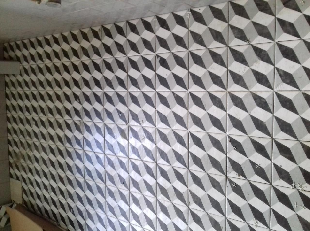 Ściana wyłożona kafelkami z geometrycznym wzorem 3D w odcieniach szarości, widoczne krzyżyki dystansowe między płytkami, surowy stan wykończenia.