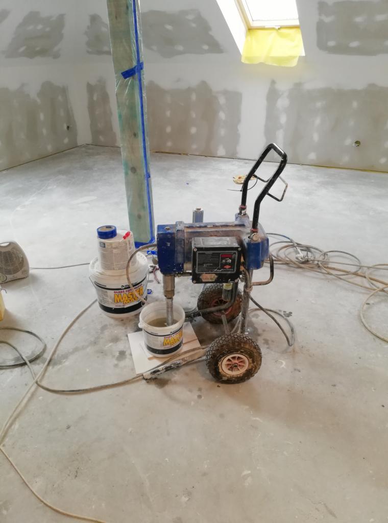 Agregat do malowania natryskowego na budowie w pokoju na poddaszu ze skośnym oknem, ściany przygotowane do malowania, zaklejona rura, widoczne wiaderka z farbą.