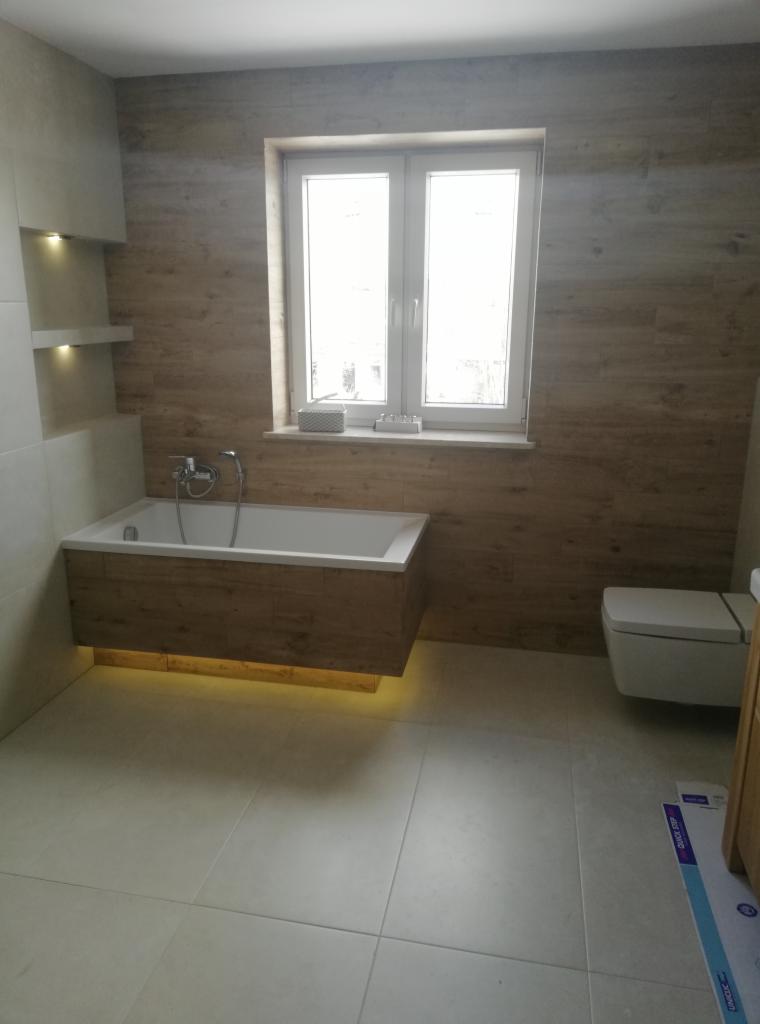 Nowoczesna łazienka z wanną podświetloną od dołu, imitującą drewno okładziną ścienną i jasnymi płytkami podłogowymi, widok na okno z dekoracjami.