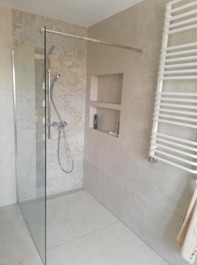 Nowoczesna kabina prysznicowa walk-in z szarymi płytkami imitującymi kamień, wbudowanymi półkami ściennymi i białym grzejnikiem drabinkowym.