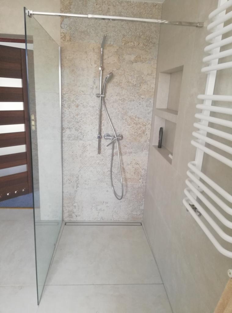 Nowoczesna kabina prysznicowa typu walk-in z odpływem liniowym, wykończona płytkami imitującymi kamień, z grzejnikiem drabinkowym i wnęką na kosmetyki.