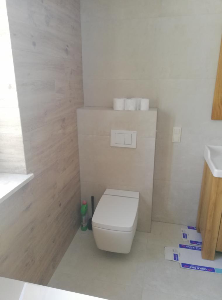 Nowoczesna toaleta podwieszana z białym panelem sterującym, na którym leżą rolki papieru toaletowego. Ściany wyłożone płytkami imitującymi drewno i gładkimi, jasnymi płytkami. Widoczne opakowania...