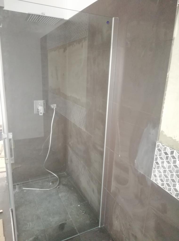 Montaż kabiny prysznicowej typu walk-in z szarymi płytkami imitującymi kamień na ścianach, widoczne niedokończone fugowanie i wystający kabel od słuchawki prysznicowej.