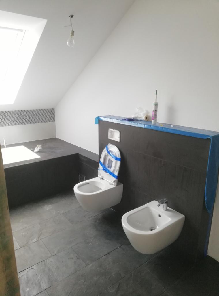 Nowoczesna łazienka na poddaszu z czarnymi płytkami podłogowymi i częściowo na ścianie, wisząca toaleta z zabezpieczeniem na desce, bidet oraz wanna pod oknem dachowym.