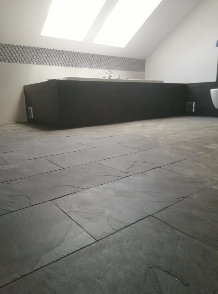 Wykończona łazienka na poddaszu ze skośnym sufitem i oknem dachowym, z czarną obudową wanny i szarymi płytkami podłogowymi imitującymi kamień.