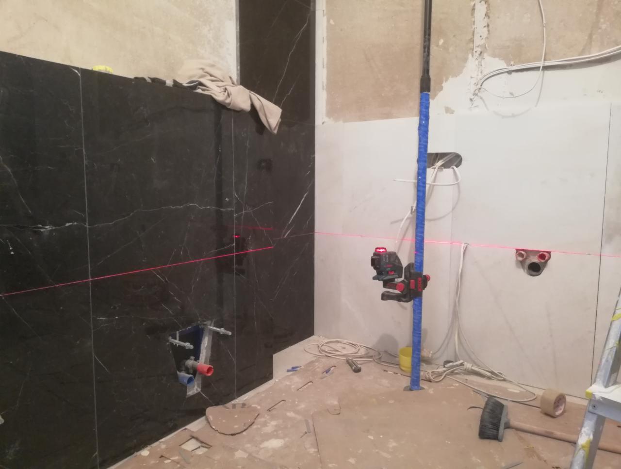Montaż czarnych, marmurkowych płytek ściennych w łazience, z widocznym laserowym niwelatorem i instalacją hydrauliczną w ścianie.
