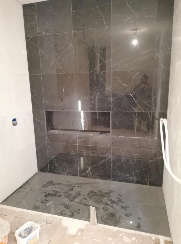 Łazienka w trakcie wykańczania: ciemne, marmurkowe płytki na ścianie z wnęką, podłoga częściowo wyłożona tymi samymi płytkami, widoczne elementy instalacji wodnej i resztki po pracach glazurniczych.