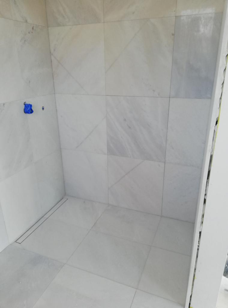 Fragment łazienki w trakcie wykańczania, widoczne białe marmurowe płytki ułożone na ścianach i podłodze, z niebieskim zabezpieczeniem na wystających rurach i liniowym odpływem podłogowym.
