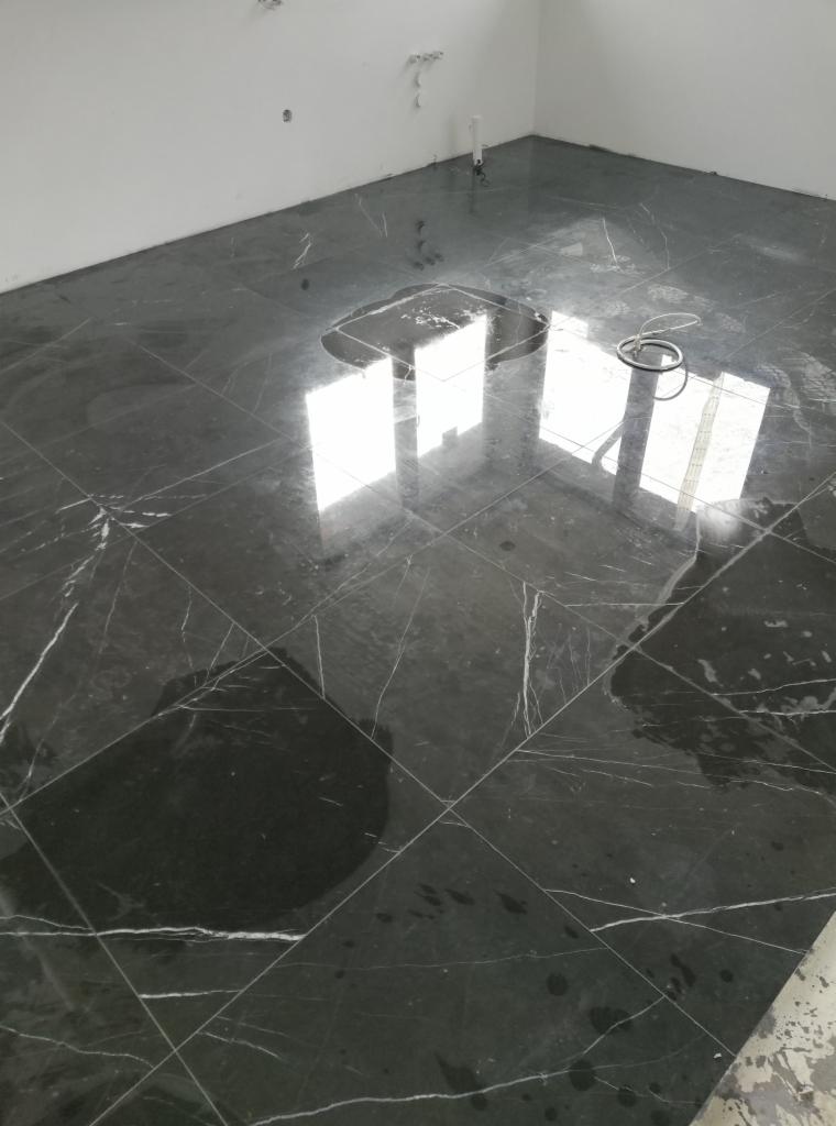 Ułożona, czarna, marmurkowa podłoga z białymi żyłami, mokra od świeżego kleju lub wody, z odbiciami okien i leżącą obok zwiniętą rurką.