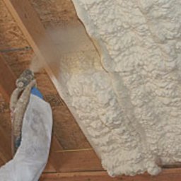 SCANDIPUR SPÓŁKA Z OGRANICZONĄ ODPOWIEDZIALNOŚCIĄ - Aplikacja izolacji pianką na poddaszu, widoczna osoba w kombinezonie ochronnym z pistoletem natryskowym i świeżo nałożona warstwa pianki.