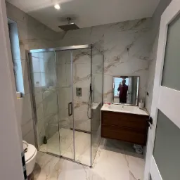 Nowoczesna łazienka po remoncie z kabiną prysznicową typu walk-in, umywalką z szafką z ciemnego drewna i marmurowymi płytkami na ścianach i podłodze. W lustrze odbija się osoba robiąca zdjęcie.