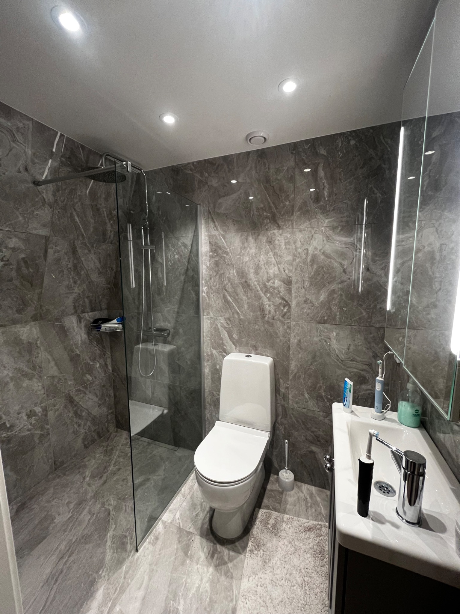 Nowoczesna łazienka z kabiną prysznicową typu walk-in, toaletą i umywalką z szafką, wykończona płytkami imitującymi marmur w odcieniach szarości.