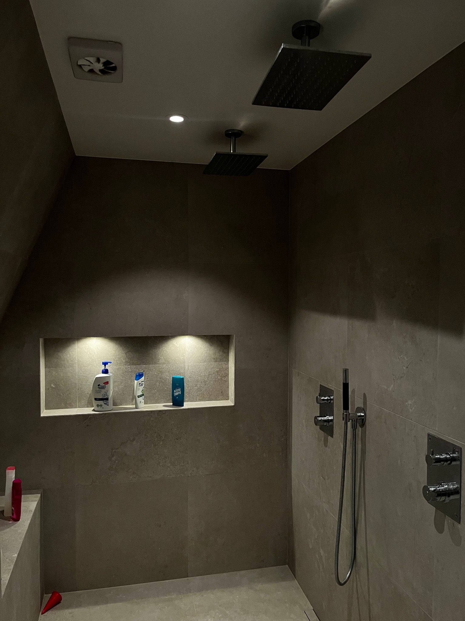 Nowoczesna łazienka z dwoma deszczownicami, wnęką z oświetleniem i kosmetykami, wykończona szarą płytką o matowej fakturze.