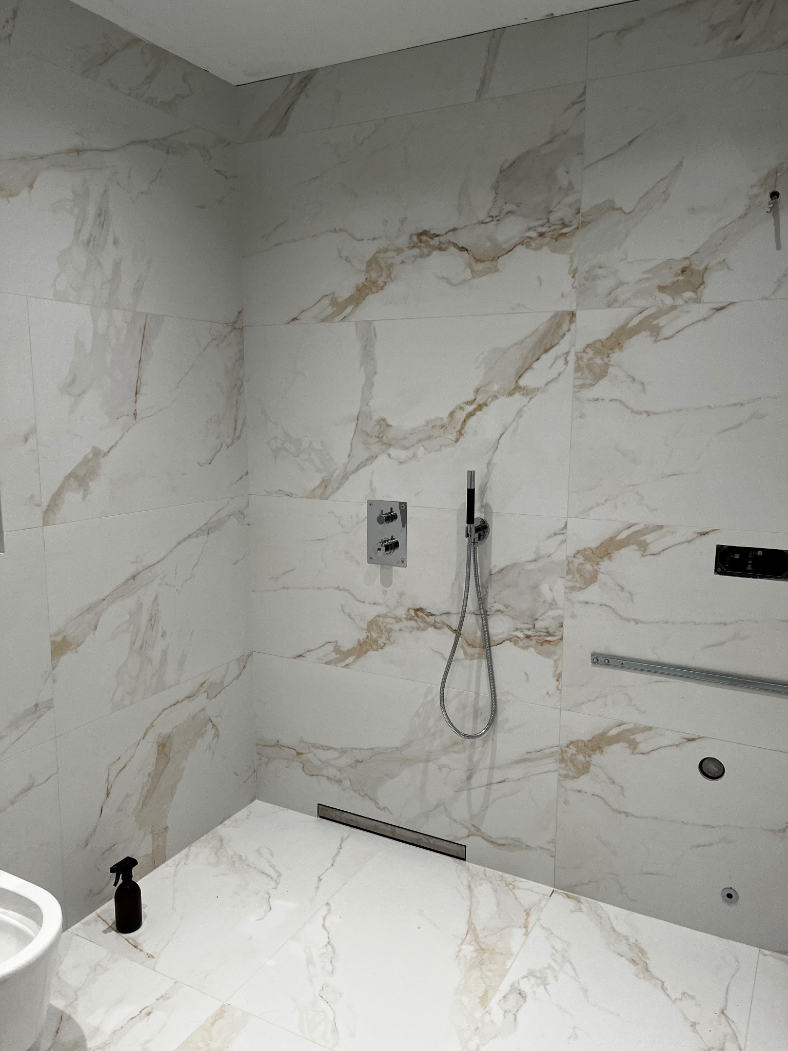 Nowoczesna łazienka w trakcie wykańczania: białe płytki imitujące marmur z brązowymi żyłami, zamontowana armatura prysznicowa, odpływ liniowy w podłodze, widoczny fragment toalety i butelka...