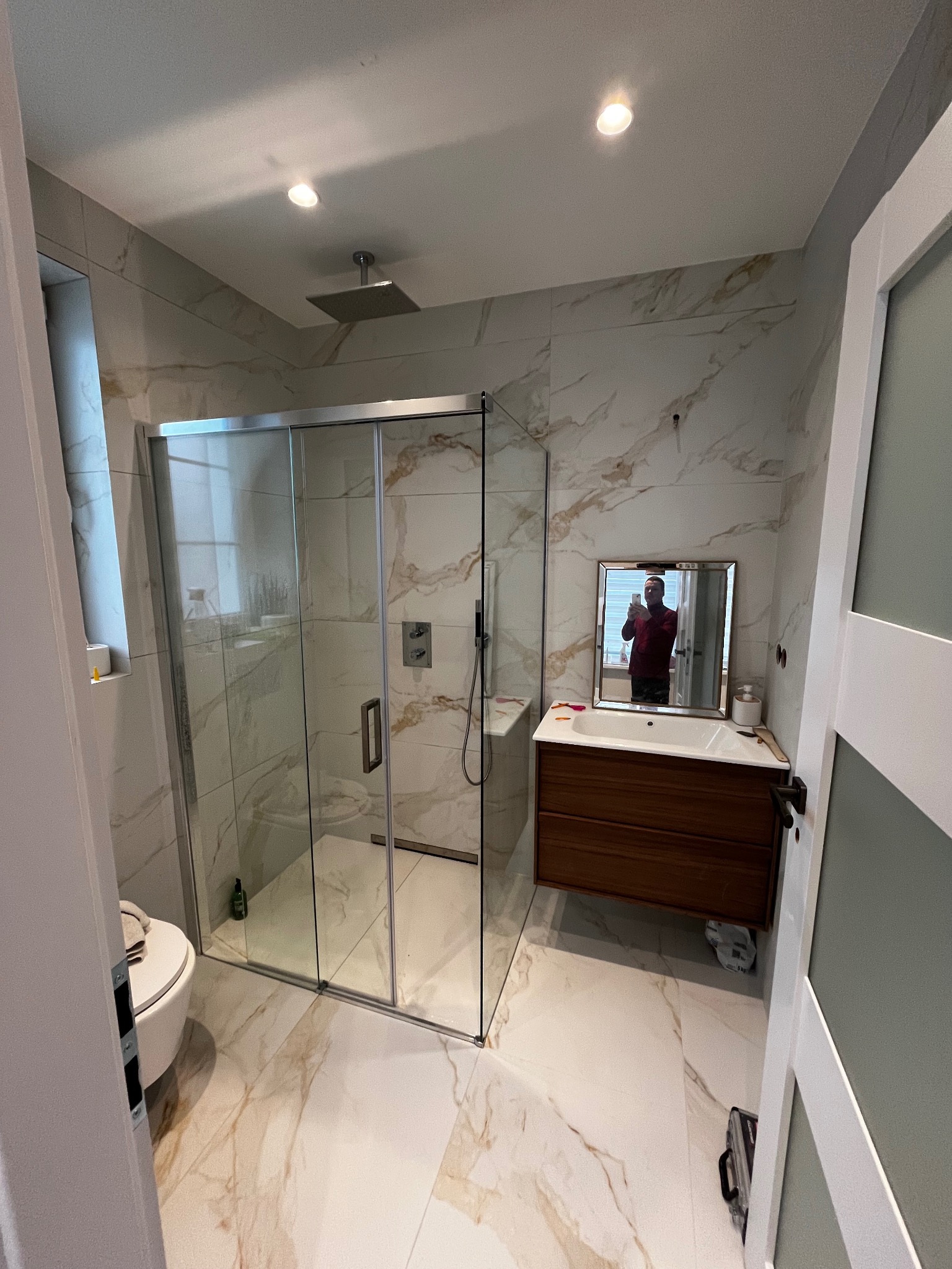 Nowoczesna łazienka po remoncie z kabiną prysznicową typu walk-in, umywalką z szafką z ciemnego drewna i marmurowymi płytkami na ścianach i podłodze. W lustrze odbija się osoba robiąca zdjęcie.