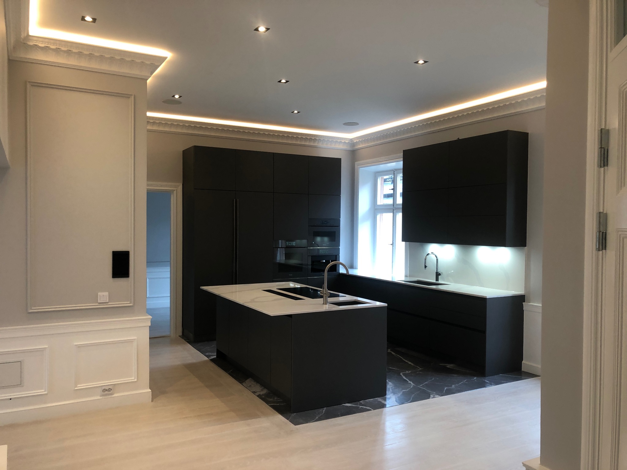Elegancka kuchnia z czarnymi szafkami, białym blatem i marmurową podłogą, oświetlenie LED podsufitowe, widok z sąsiedniego pomieszczenia przez otwarte drzwi.