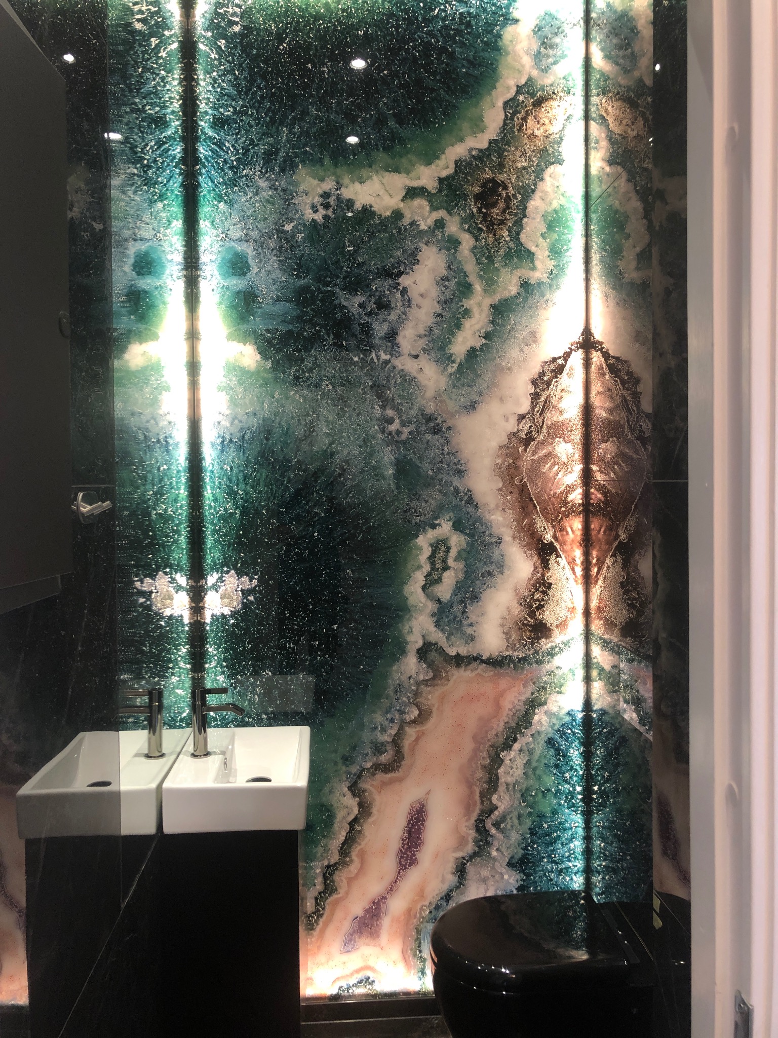 Luksusowa łazienka z podświetlaną ścianą z onyksu, dwie białe umywalki na czarnym blacie i czarna toaleta, elegancki design.