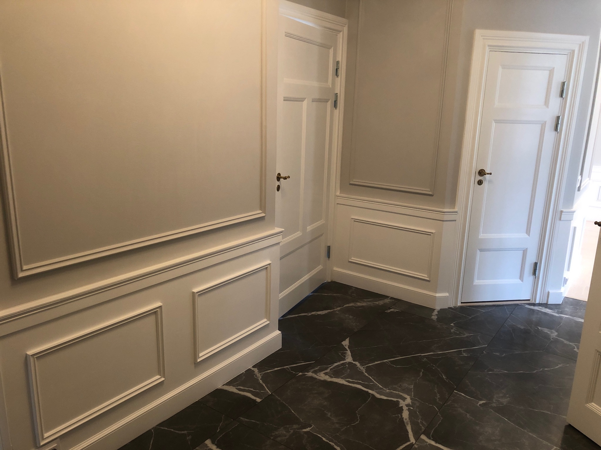 Wykończony korytarz w Bielkowie z szarymi ścianami zdobionymi sztukaterią, białymi drzwiami z mosiężnymi klamkami i podłogą z ciemnych płytek imitujących marmur.