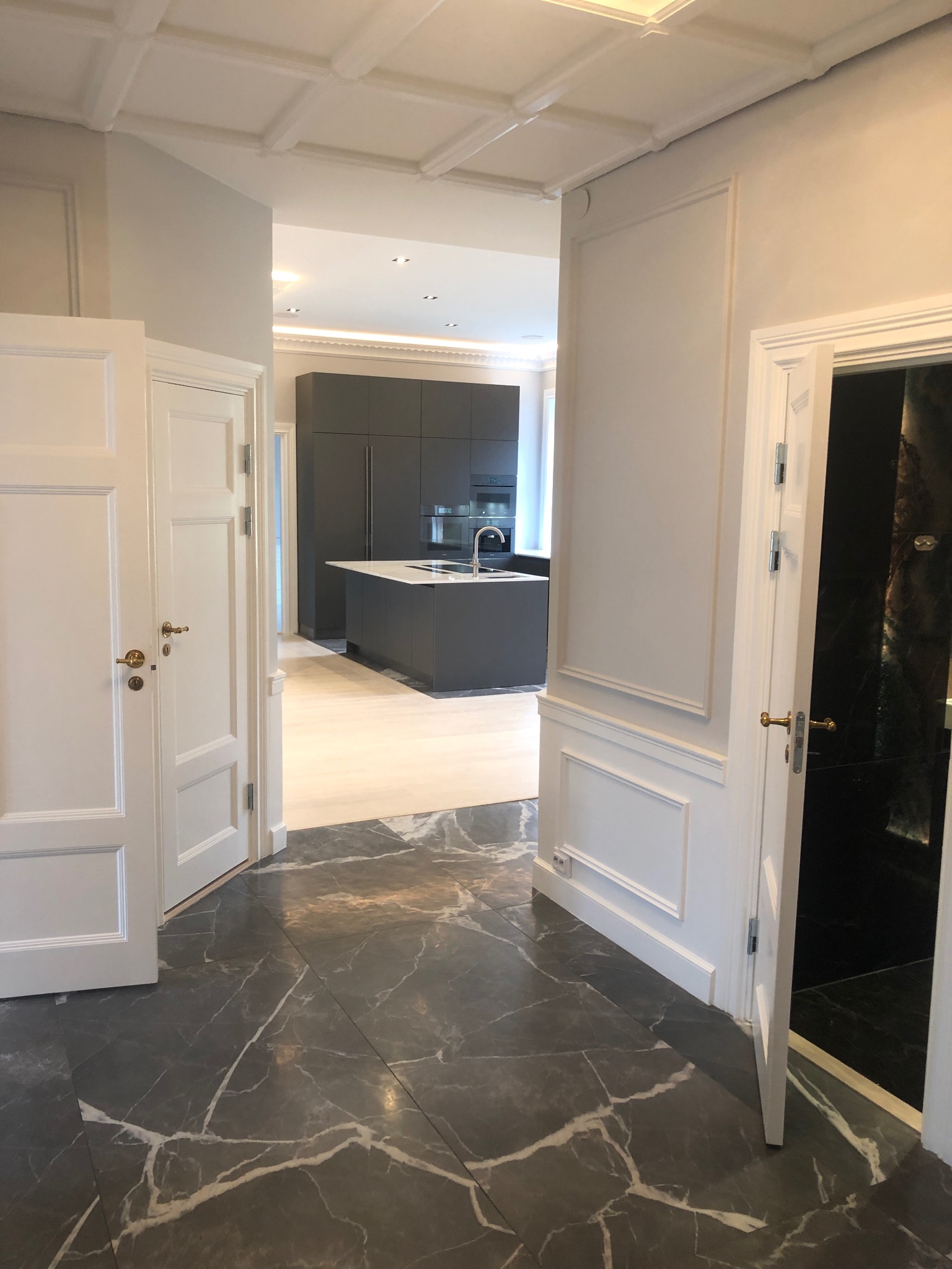 Eleganckie wnętrze z otwartą kuchnią, widok z korytarza z marmurową podłogą i białymi drzwiami z ozdobnymi listwami.