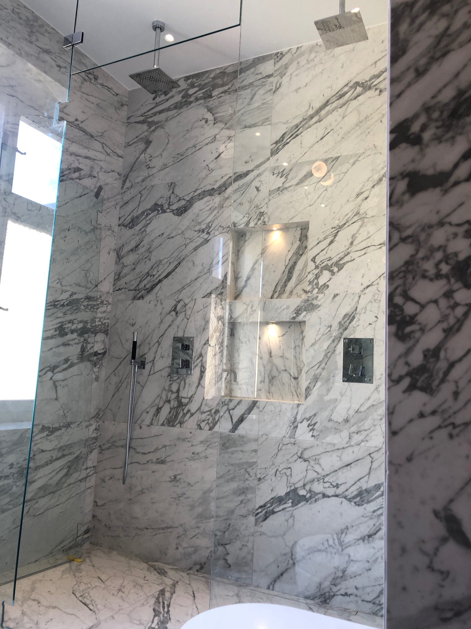 Luksusowa kabina prysznicowa walk-in wykończona białym marmurem z szarymi żyłkami, wyposażona w dwa deszczownice i wnęki z oświetleniem punktowym.