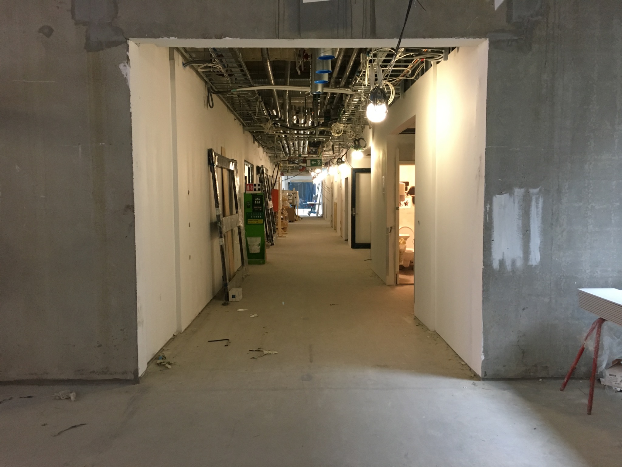Długi korytarz w surowym stanie, z widocznymi instalacjami pod sufitem i przygotowanymi do montażu ościeżnicami, ściany częściowo otynkowane, widoczne ślady prac budowlanych.