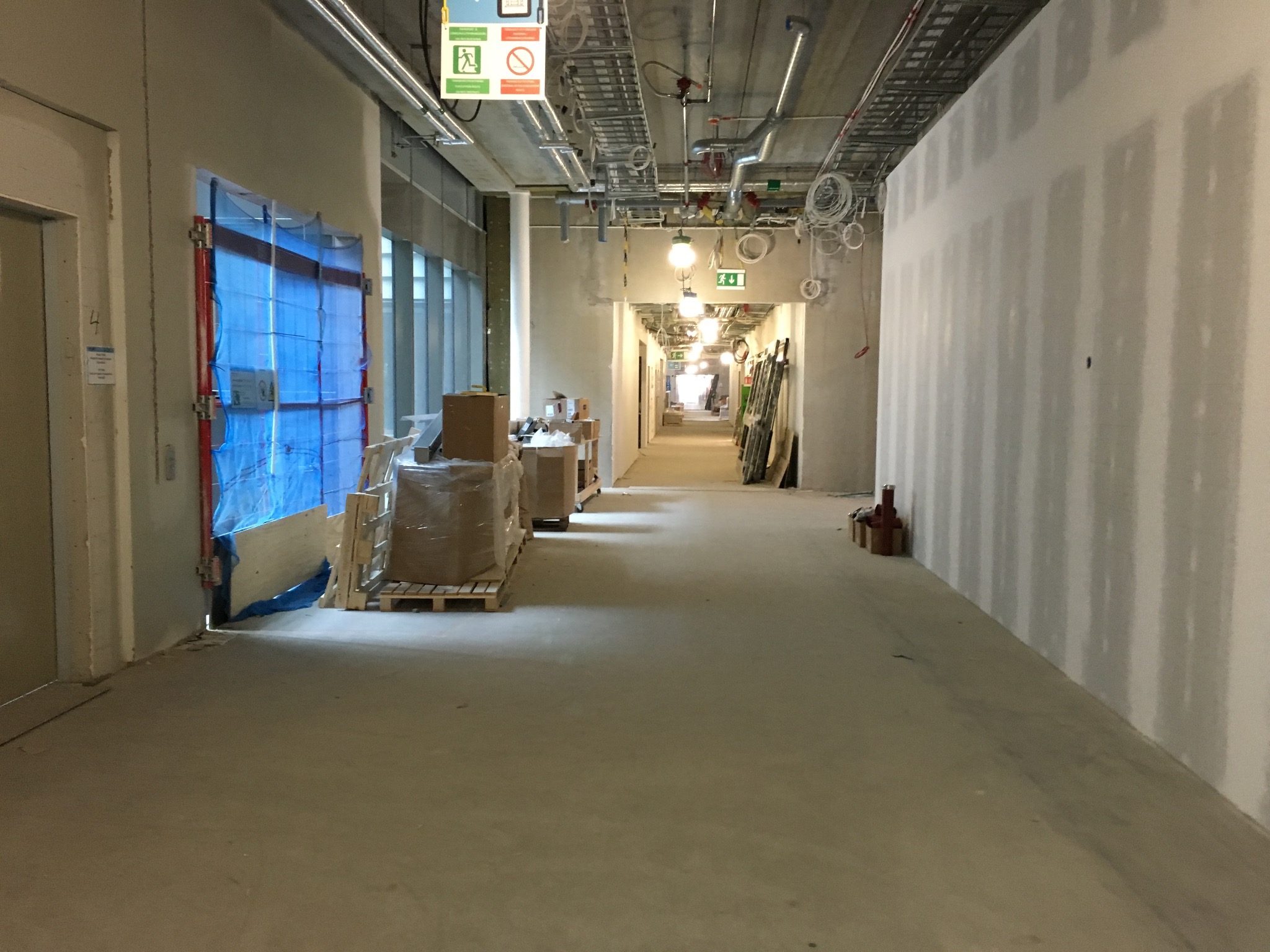 Korytarz w trakcie prac wykończeniowych w budynku biurowym, widoczne instalacje pod sufitem, ściany przygotowane pod malowanie, zabezpieczone okna i materiały budowlane.