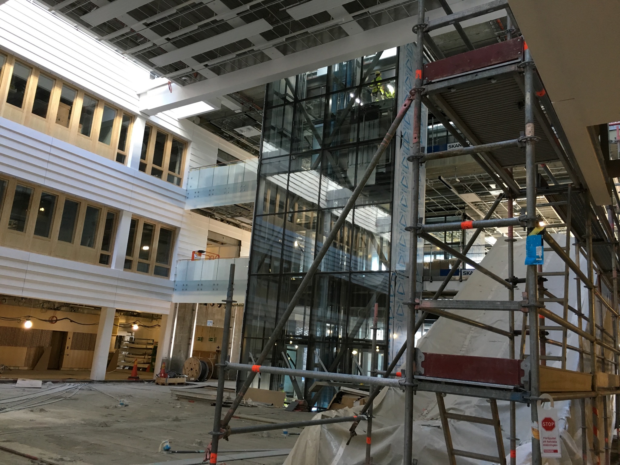 Wnętrze budynku w trakcie prac wykończeniowych, widoczne rusztowania, szklana konstrukcja windy i pracownicy w oddali.