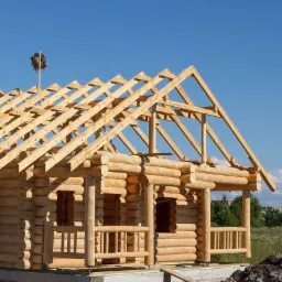 Konstrukcja drewnianego domu z bali w trakcie budowy, widoczne krokwie dachowe i wieniec na szczycie, błękitne niebo w tle.