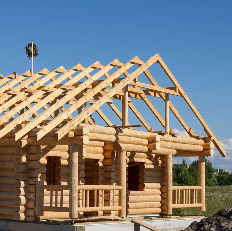 Konstrukcja drewnianego domu z bali w trakcie budowy, widoczne krokwie dachowe i wieniec na szczycie, błękitne niebo w tle.