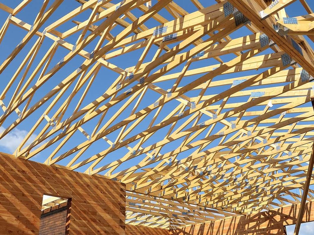 Konstrukcja drewniana dachu z widocznymi wiązarami kratowymi i łącznikami metalowymi na tle błękitnego nieba, widok od wewnątrz budynku w trakcie budowy.