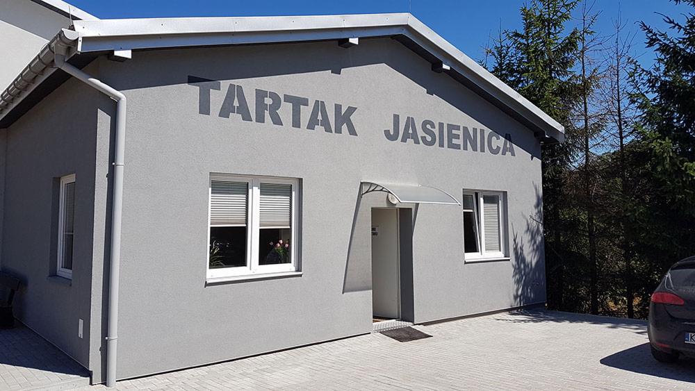 Budynek tartaku w Jasienicy z szarymi ścianami, dwoma oknami z żaluzjami i napisem 'TARTAK JASIENICA' nad wejściem.