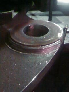 Spoina spawalnicza wokół metalowego otworu na ciemnej, metalowej powierzchni. Widoczne ślady ciepła i niedoskonałości spoiny.