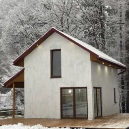 Nowoczesny dom jednorodzinny z drewnianym tarasem, pokryty śniegiem, na tle zasypanego śniegiem lasu.