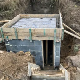 Budowa zejścia do piwnicy z betonowych bloczków, widoczne drewniane szalunki i zbrojenie, ziemia wokół wykopu.