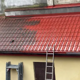 Częściowo wyczyszczony czerwony dach z metalowej dachówki, widoczne różnice w kolorze, drabina oparta o ścianę budynku, złożone rusztowanie w tle.