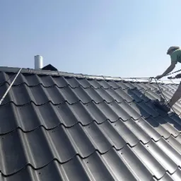 Pracownik z zabezpieczeniem linowym maluje ciemnoszary dach z blachodachówki modułowej, widoczny komin i miarka zwisająca z krawędzi dachu.