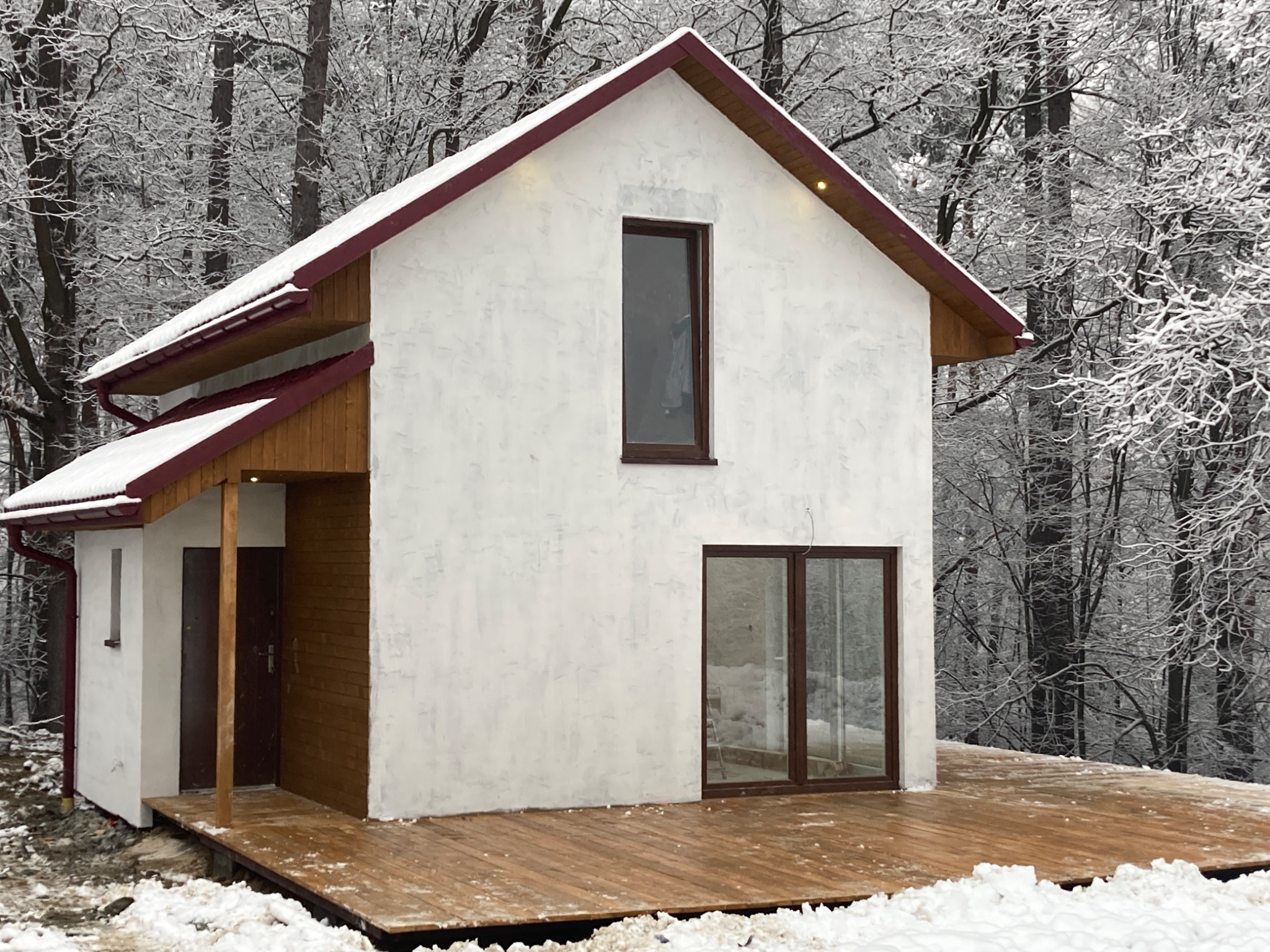 Nowoczesny dom z białym tynkiem i bordowym dachem, otoczony zaśnieżonym lasem, z widocznym drewnianym tarasem i częściowo odśnieżonym dachem.