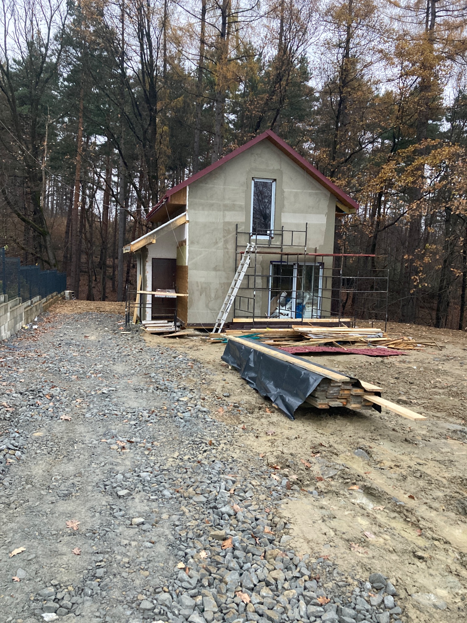 Dom w trakcie budowy z częściowo wykończonym dachem, rusztowaniami i stosem materiałów budowlanych na ziemi. Widoczne drzewa w tle.