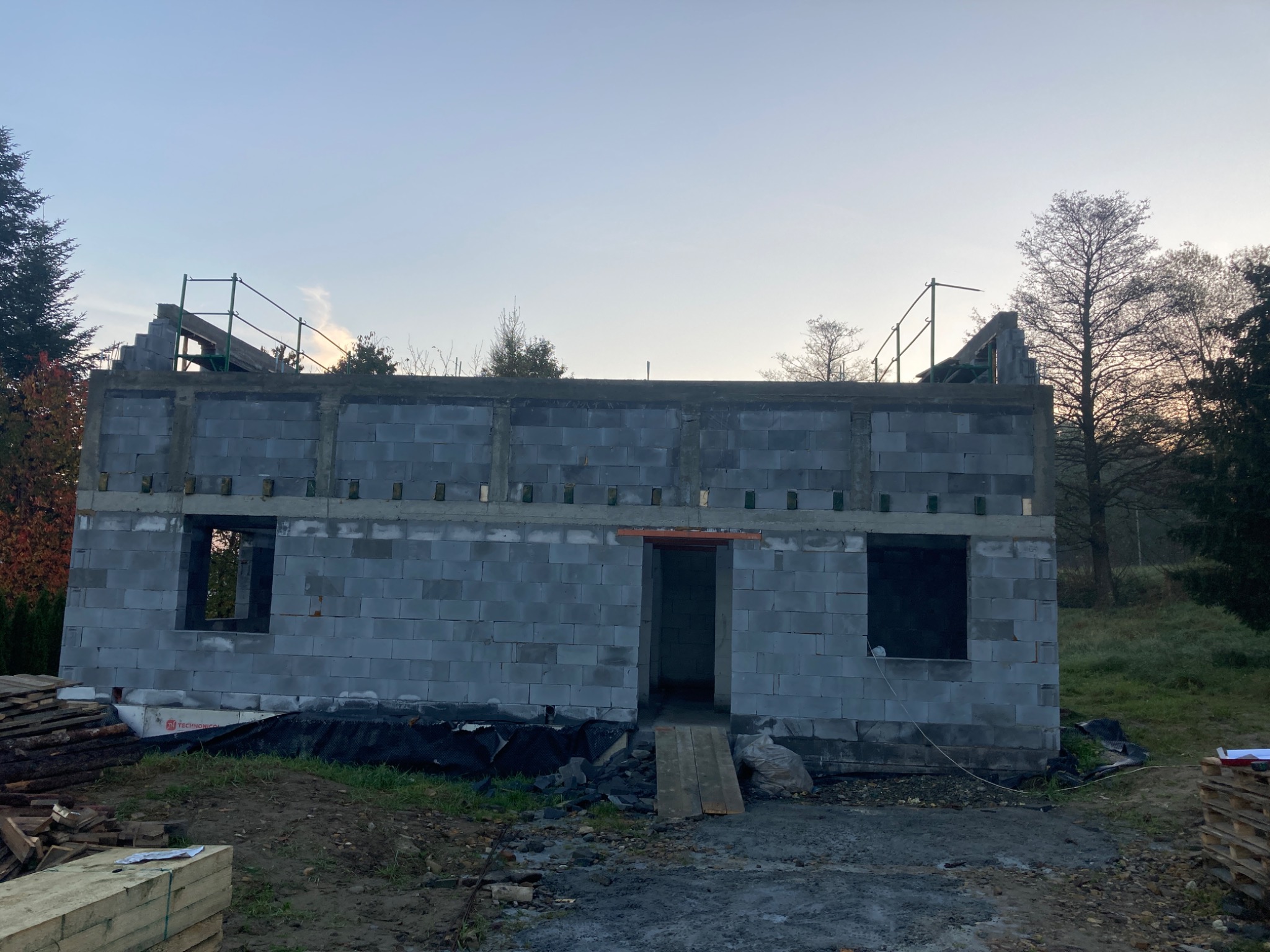 Surowy budynek z szarej cegły w trakcie budowy, widoczne otwory okienne i drzwiowe, na dachu rusztowanie, w tle drzewa i niebo.