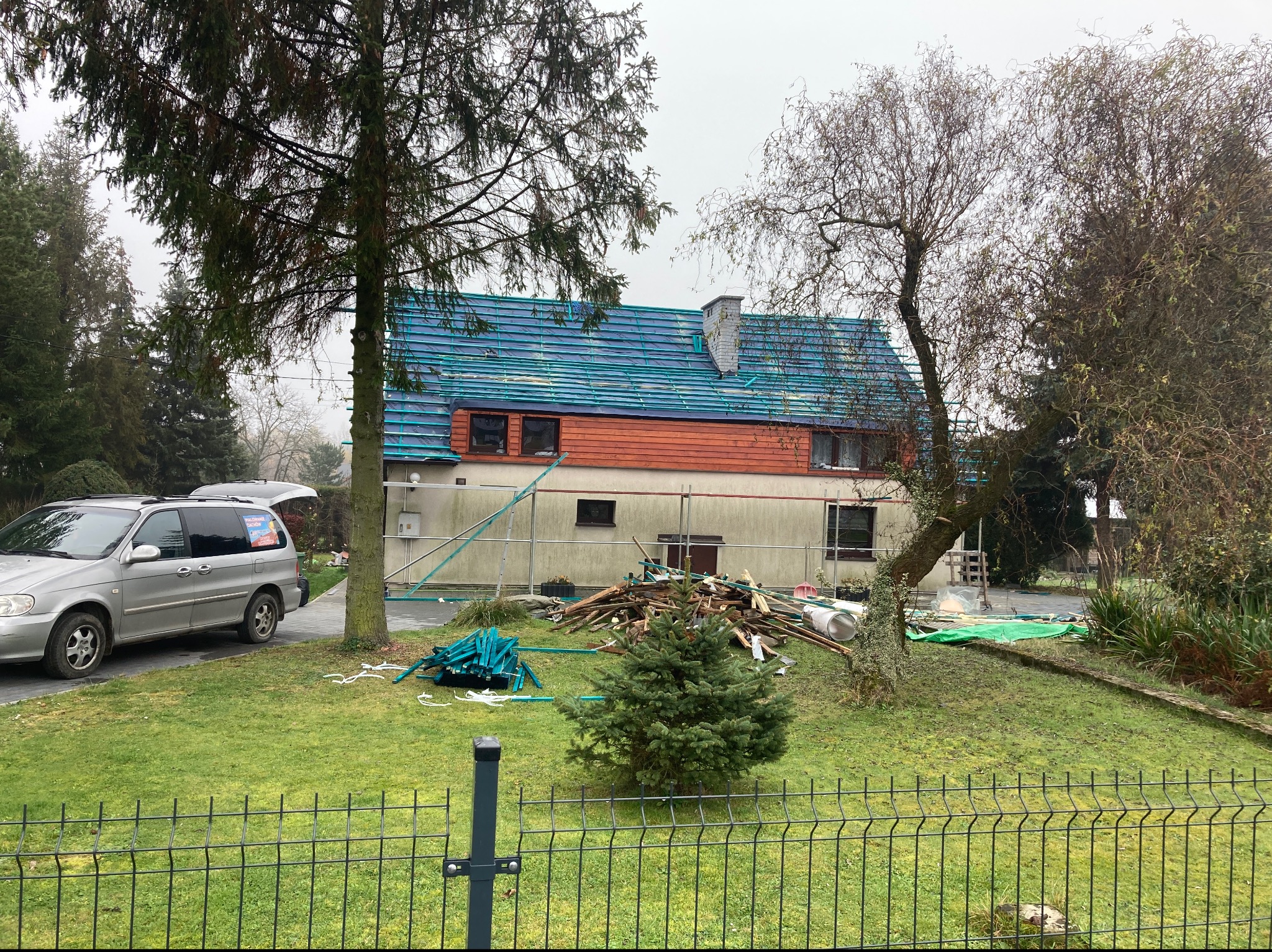 Dom jednorodzinny z częściowo wymienionym dachem; widoczne zabezpieczenie folią, nowe niebieskie dachówki, drewniane elementy elewacji, rusztowanie oraz sterta odpadów po pracach dekarskich...