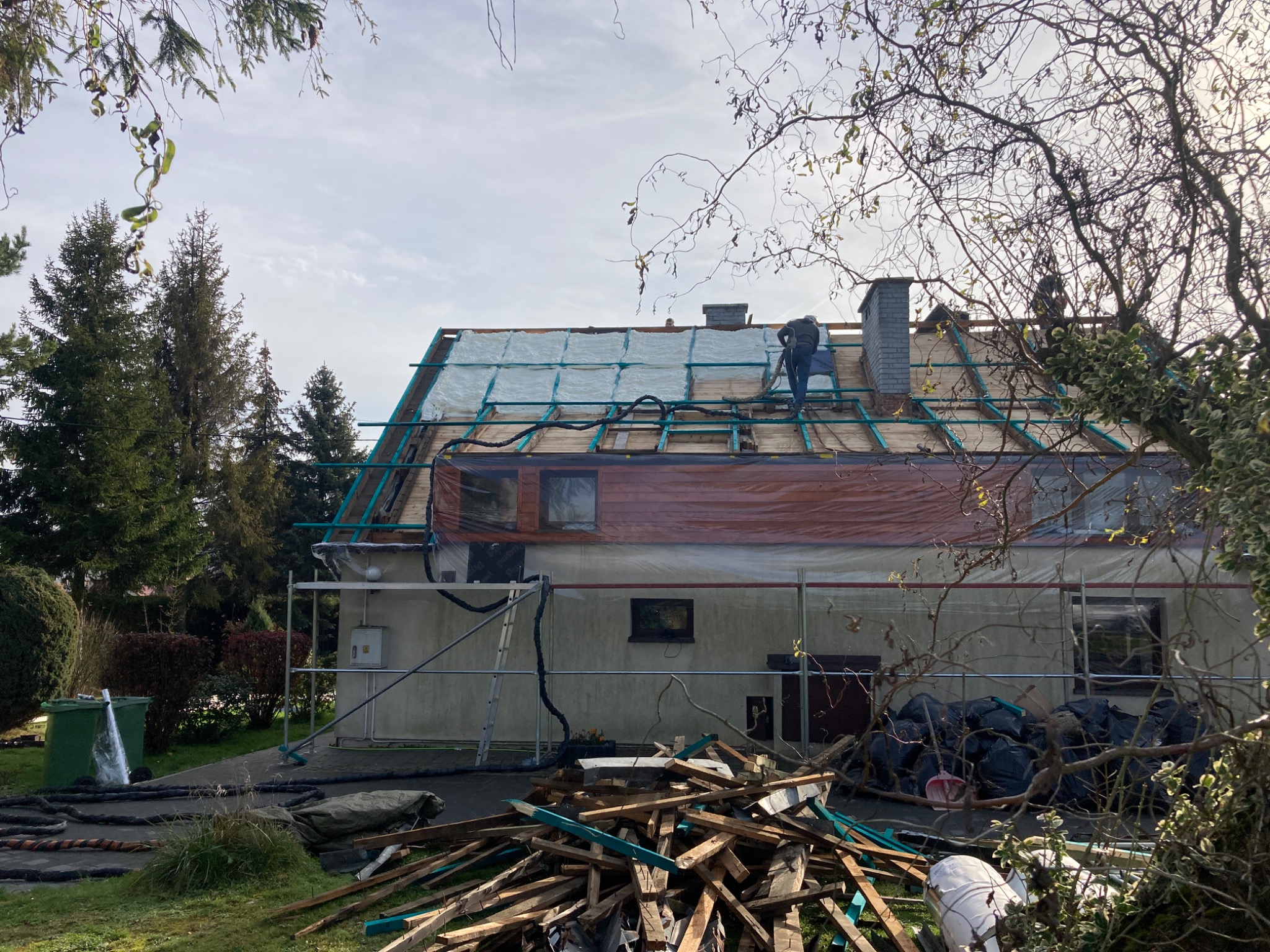 Pracownik na dachu domu jednorodzinnego, widoczny na tle drzew, wykonujący prace związane z izolacją termiczną, na dachu rozłożona biała izolacja i folia ochronna, w tle komin z cegły.