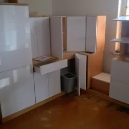 Montaż białej zabudowy kuchennej z drewnianymi elementami, widoczna wysunięta szuflada z dokumentami i kosz na śmieci w przestrzeni szafki pod blatem.