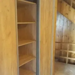 Otwarta szafa wnękowa z jasnego drewna z przesuwnymi drzwiami, widok na wnętrze z półkami, w tle drewniana konstrukcja antresoli pod skośnym dachem.