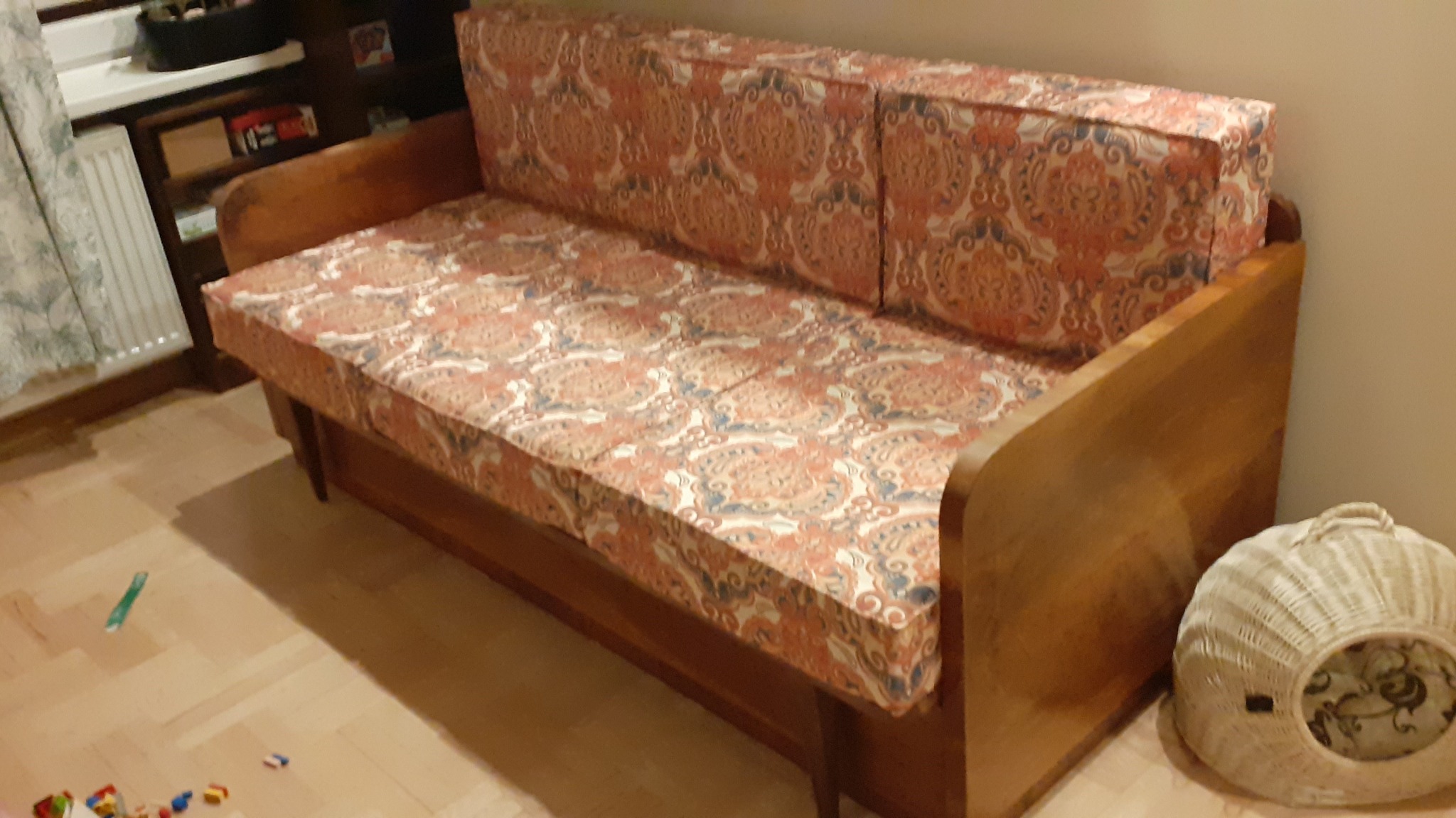 Rozkładana sofa z drewnianymi bokami i wzorzystą tapicerką w odcieniach czerwieni i błękitu, obok wiklinowa budka dla kota.