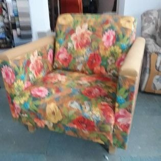 Fotel tapicerowany z wzorem kwiatowym, beżowe podłokietniki, widoczne niedoskonałości tapicerki i tło sugerujące sklep lub magazyn.