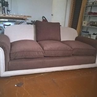 Trzyosobowa kanapa z beżowym obiciem ramy i brązowymi poduszkami siedziskowymi oraz oparciową, widoczna w warsztacie stolarskim z maszynami w tle.